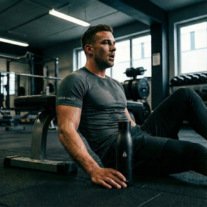 Man sitting on a bench in a gym holding a shadow black AQUA VAULT PureX Smart Water Bottle.