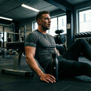 Man sitting on a bench in a gym holding a black AQUA VAULT PureX Smart Water Bottle