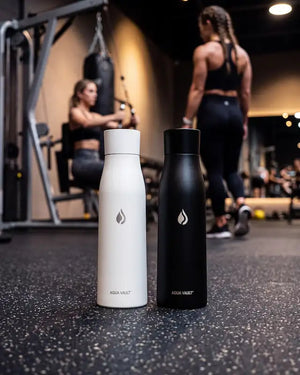 AQUA VAULT PureX UV-C self-cleaning smart water bottle, one matte white and one matte black, standing side-by-side on a gym floor with blurred gym-goers in the background.